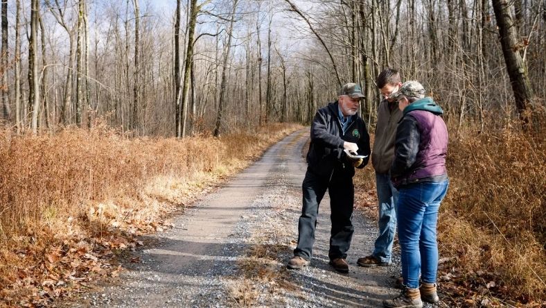 Three people standing on rural path reviewing notes in wooded landscape